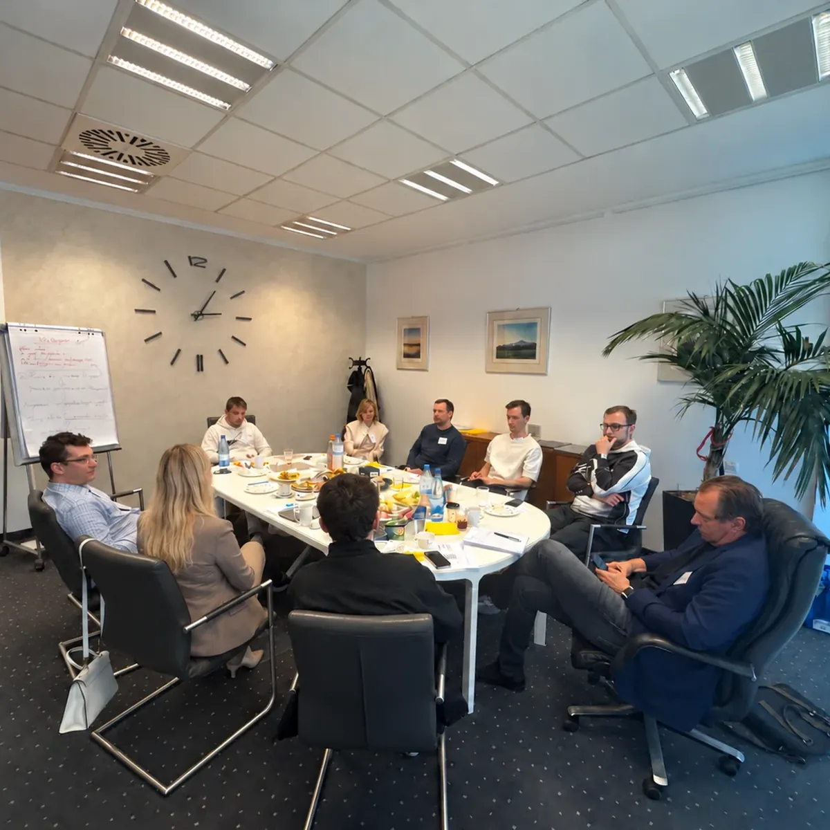 Entrepreneurs seated around a workshop table in a meeting room with a light textured wall and a large clock